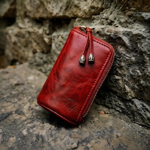May include: A rectangular red leather key case with a wrap-around zipper closure. The case features a textured surface, a debossed logo on the lower front, and two leather zipper pulls with decorative metallic end caps in a bronze finish.