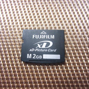 Peut inclure: Une carte mémoire Fujifilm xD-Picture Card noire avec du texte blanc indiquant FUJIFILM, xD, xD-Picture Card et M 2GB. La carte rectangulaire présente une petite icône de flèche triangulaire en haut au centre et une petite zone d'étiquette.