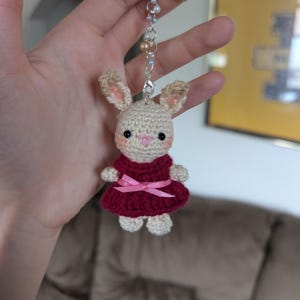 Op de afbeelding: Een kleine gehaakte konijn-sleutelhanger met een crèmekleurig lichaam, roze wangen en een donkerrode jurk met een lichtroze strik. Het konijn is bevestigd aan een zilverkleurige metalen ketting versierd met kleine iriserende en perzikkleurige kralen.