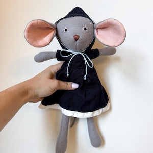 May include: A plush mouse toy with grey fabric skin, large pink-lined ears, and embroidered facial features. The mouse wears a black hooded dress with a white hem and a thin light blue ribbon tied in a bow at the neckline, featuring long arms and legs.