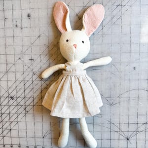 May include: A stuffed rabbit toy with cream-colored fabric, long ears with pink inner linings, and a small pink embroidered nose. The rabbit wears a sleeveless, gathered beige dress with a simple bodice and a full, pleated skirt reaching its waist.