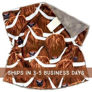 May include: A brown and white patterned blanket featuring a repeating design of Highland cow faces. The blanket has a soft, gray, plush backing. The design includes the text "SHIPS IN 3-5 BUSINESS DAYS".