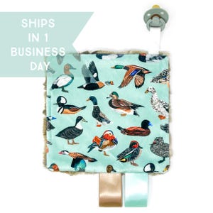 May include: A baby security blanket with a duck pattern on a light blue background. The blanket has a soft, plush border and attached ribbons. A pacifier is attached to the top of the blanket. The text "SHIPS IN 1 BUSINESS DAY" is in the upper left corner.