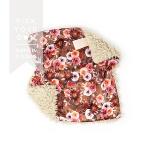 May include: A floral patterned blanket with a brown background and a cream-colored, fluffy trim. The blanket features a repeating pattern of pink, red, and orange roses. The text "PICK YOUR OWN" and "SHIPS IN 3-5 DAYS" is visible.