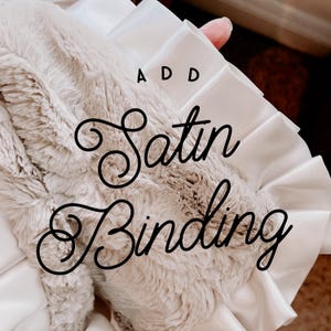 May include: A plush, beige blanket with a white satin binding. The words "ADD Satin Binding" are written in a decorative script across the blanket. The blanket has a soft, furry texture.