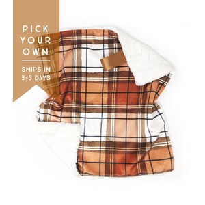 May include: A plaid blanket in shades of brown, orange, and white, with a soft, white, plush lining. The blanket has a rectangular shape and a satin ribbon tag. The text "PICK YOUR OWN" and "SHIPS IN 3-5 DAYS" is visible.