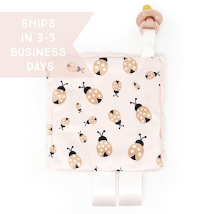 May include: A baby pacifier with a pink and white ladybug print security blanket. The blanket has a soft, plush border and white ribbon attachments. The pacifier is light pink with a yellow nipple. The text "SHIPS IN 3-5 BUSINESS DAYS" is visible.