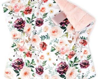 May include: A floral baby blanket with a white background and a pattern of pink, red, and purple roses. The blanket has a soft, pink, plush backing. A pink ribbon is attached to the blanket.