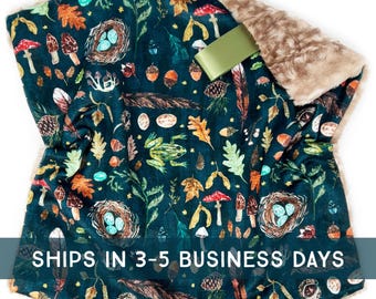 May include: A teal blanket with a woodland pattern featuring mushrooms, leaves, acorns, and a frog. The blanket has a soft, faux fur trim and a green ribbon. The text "SHIPS IN 3-5 BUSINESS DAYS" is printed on the blanket.