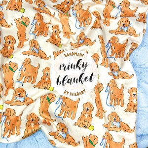 May include: A cream-colored blanket featuring a repeating pattern of golden retriever illustrations with blue leashes and bandanas. The blanket has a blue plush border and the text "Handmade minky blanket by iviebaby".