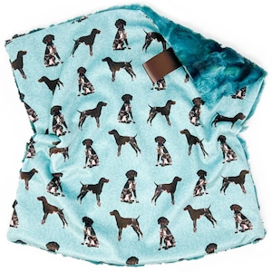 May include: A light blue blanket featuring a repeating pattern of German Shorthaired Pointers in various poses. The blanket has a teal faux fur trim and a brown leather tag. The blanket is made of soft material.