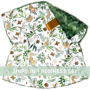 May include: A white baby blanket with a woodland animal print featuring foxes, owls, squirrels, and rabbits. The blanket has a green plush backing and a gold satin ribbon. The blanket is a soft, cozy item.