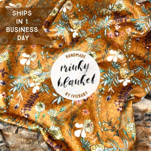 May include: A handmade minky blanket with a floral pattern in shades of gold, white, yellow, and teal. The blanket features the text "HANDMADE minky blanket BY IVIEBABY" in a circular design. The blanket is soft and plush.