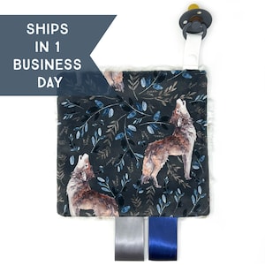 May include: A soft, plush baby toy with a dark blue and gray wolf print. The toy has a white faux fur border and two satin ribbons in gray and blue. A pacifier clip is attached to the top.