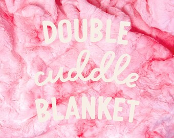 May include: A plush, pink blanket with a textured surface. The words "DOUBLE cuddle BLANKET" are written across the blanket in a cream-colored font. The blanket appears soft and cozy.