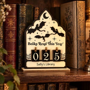 Puede incluir: Un rastreador de libros de madera color crema con una parte superior en arco apuntado, con siluetas negras de tres dragones voladores, estrellas y una luna creciente. Muestra el texto "Books Read This Year" y "Sally's Library" con tres tarjetas numéricas negras.
