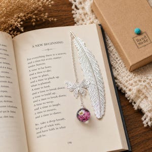 Puede incluir: Un marcapáginas de metal plateado con un diseño detallado de pluma, un delicado dije de mariposa y una pequeña esfera de cristal que contiene diminutas flores rosas y blancas. El marcapáginas descansa sobre una página de libro titulada A New Beginning.