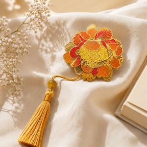 Puede incluir: Un marcapáginas de metal dorado con un diseño de flor grande e intrincado con pétalos en capas de esmalte amarillo, naranja y rojo. Un cordón dorado conecta la flor a una borla de seda larga de color amarillo dorado con una pequeña cuenta.