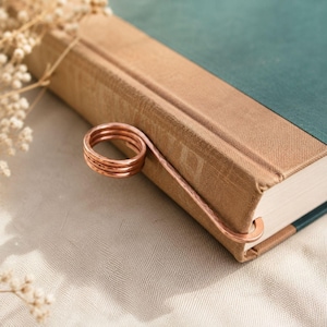 May include: A copper-colored metal page holder featuring a coiled ring design attached to a flat, curved arm that slides over the spine of a closed book with a tan fabric cover and a dark green accent panel on the front cover.