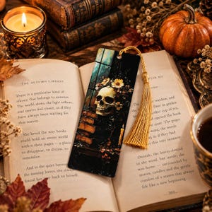 Puede incluir: Un marcapáginas rectangular con una ilustración oscura de un cráneo humano adornado con flores amarillas, junto a una pila de libros y una vela encendida, rematado con una larga borla de color oro metálico en el orificio superior.