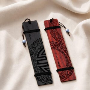 Puede incluir: Dos marcapáginas rectangulares de madera, uno negro y otro marrón rojizo, con intrincados patrones grabados con láser, bandas elásticas negras y un cordón marrón con una pequeña cuenta de cerámica con motivos florales azules y blancos.