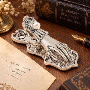 May include: A silver-toned metal clip featuring a sculpted hand design with a white pearl accent at the wrist. The clip has an ornate, textured base and a spring mechanism. The hand is positioned as if resting on the base, which has a decorative border.