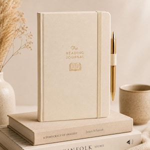 Puede incluir: Un diario de lectura de tapa dura color crema con texto dorado que dice The Reading Journal y un icono de libro. Una banda elástica sujeta un bolígrafo de metal dorado en el lateral. El diario descansa sobre dos libros apilados con cubiertas neutras.