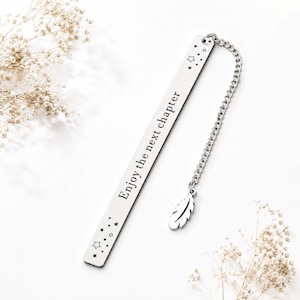 Puede incluir: Un marcapáginas de metal plateado de forma rectangular, grabado con el texto "Enjoy the next chapter" y motivos de estrellas pequeñas. Una cadena plateada está unida a la parte superior, con un pequeño dije de pluma de metal colgando.