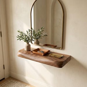 Könnte beinhalten: Ein wandmontiertes, schwebendes Regal aus dunklem Holz mit natürlicher Baumkante, darauf eine beige Keramikvase mit grünem Blattwerk, eine hellbraune Lederablage mit Schlüsseln und ein kleines rechteckiges Tablett aus goldfarbenem Metall.