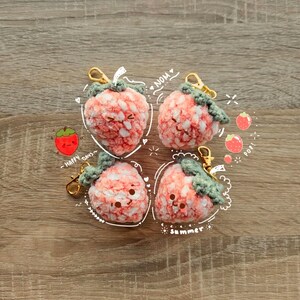 May include: Four handmade crochet strawberry keychains with gold-coloured clasps. The strawberries are pink and white with green tops. The image includes the words "NOM", "Happy Days", and "Summer".