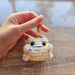 Puede incluir: Llavero artesanal con forma de nube sonriente. La nube está hecha de hilo suave multicolor en tonos amarillos, rosas, azules y blancos. Tiene un cierre metálico dorado para sujetarlo a llaves o a un bolso.