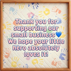 Peut inclure: Un cadre en bois carr&eacute; contenant un panneau en tissu matelass&eacute; avec un motif d'empreintes de mains multicolores. Le texte indique : Thank you for supporting our small business. We hope your little hero absolutely loves it! Un c&oelig;ur bleu est pr&eacute;sent.