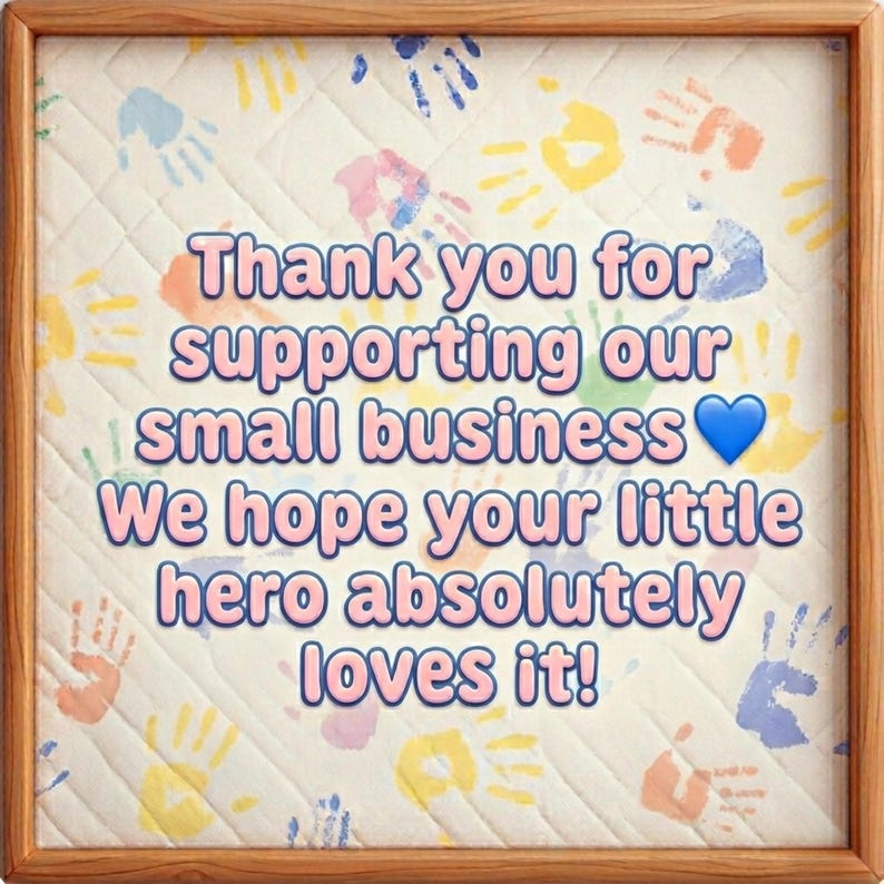 Peut inclure: Un cadre en bois carr&eacute; contenant un panneau en tissu matelass&eacute; avec un motif d'empreintes de mains multicolores. Le texte indique : Thank you for supporting our small business. We hope your little hero absolutely loves it! Un c&oelig;ur bleu est pr&eacute;sent.