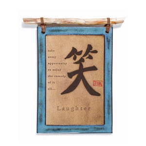 May include: A blue framed ceramic tile with a tan background. The tile features a black Japanese character for laughter, with the word "Laughter" written below it. The tile also includes a quote: "Take every opportunity to enjoy the comedy of it all..."