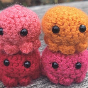 Puede incluir: Cuatro pequeñas figuras de pulpo tejidas a ganchillo, redondas, apiladas en dos columnas. La superior izquierda es rosa claro, la inferior izquierda es rojo oscuro, la superior derecha es naranja y la inferior derecha es magenta.