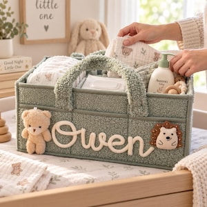 Puede incluir: Un organizador de guardería de tela sherpa verde salvia con el nombre Owen en letras cursivas blancas, un pequeño oso de peluche y un aplique de erizo. El cesto contiene artículos esenciales para bebés, incluyendo paños de muselina y loción.