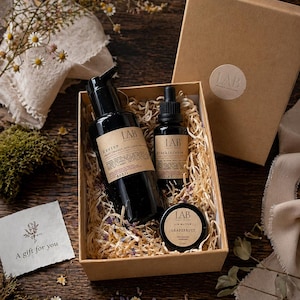 May include: A brown gift box filled with beauty products, including black bottles and a small jar. The box contains a bottle with a pump, a dropper bottle, and lip butter. The products are labelled "LAB" and are surrounded by decorative straw.