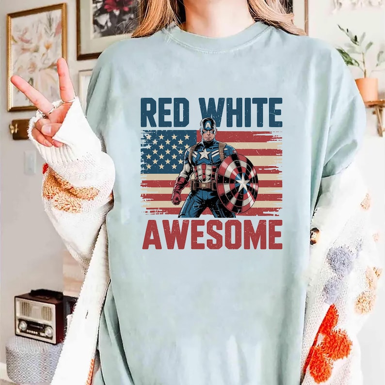 May include: A light blue t-shirt featuring a graphic of Captain America standing in front of a distressed American flag. The text "RED WHITE" appears in dark blue above the character, and the word "AWESOME" is printed in red below the graphic.