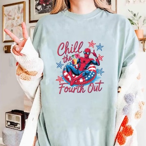 May include: A light blue t-shirt featuring a graphic of Spider-Man relaxing in a red, white, and blue inner tube while holding a drink. The text Chill the Fourth Out is printed in red, surrounded by red and blue stars on a light blue background.