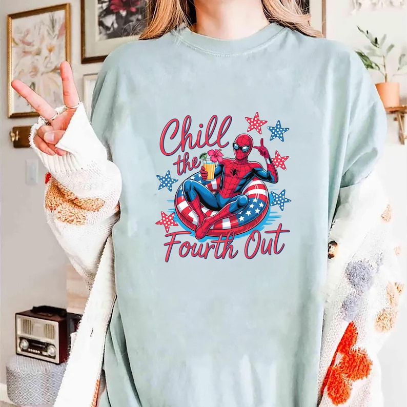 May include: A light blue t-shirt featuring a graphic of Spider-Man relaxing in a red, white, and blue inner tube while holding a drink. The text Chill the Fourth Out is printed in red, surrounded by red and blue stars on a light blue background.