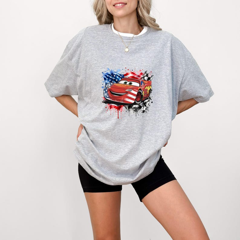 May include: A heather gray short-sleeve t-shirt featuring a graphic print of a red race car character with a smiling face. The car is overlaid with a patriotic American flag pattern, surrounded by watercolor-style blue, red, and black paint splatters.