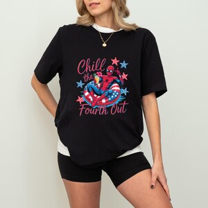 May include: A black short-sleeved t-shirt featuring a graphic of a superhero in a red and blue suit relaxing in a red, white, and blue striped pool float. The text Chill the Fourth Out is printed in pink, surrounded by red and blue star icons.