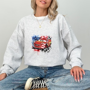 May include: A light gray crewneck sweatshirt featuring a graphic print of a red race car with a smiling face. The car is decorated with American flag patterns and surrounded by black and white checkered flag accents and colorful paint splatters.