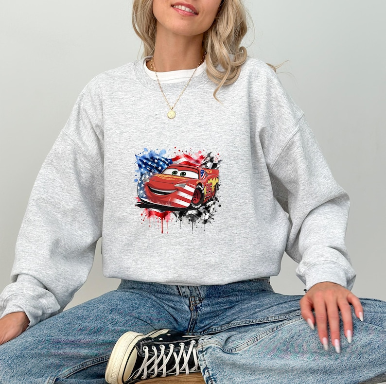 May include: A light gray crewneck sweatshirt featuring a graphic print of a red race car with a smiling face. The car is decorated with American flag patterns and surrounded by black and white checkered flag accents and colorful paint splatters.