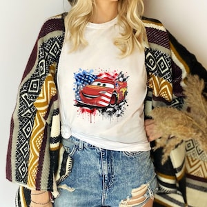 May include: A white t-shirt featuring a graphic of a red race car with American flag patterns, layered under a patterned open-front cardigan with geometric designs in black, white, yellow, and maroon, paired with distressed blue denim shorts.