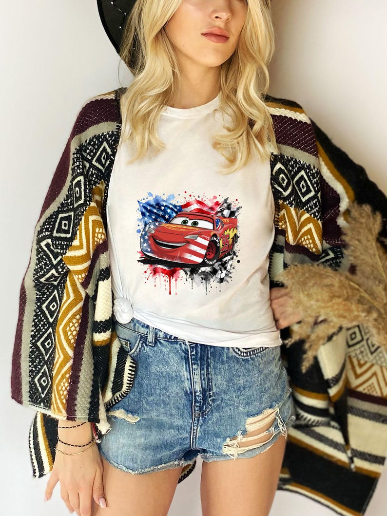 May include: A white t-shirt featuring a graphic of a red race car with American flag patterns, layered under a patterned open-front cardigan with geometric designs in black, white, yellow, and maroon, paired with distressed blue denim shorts.
