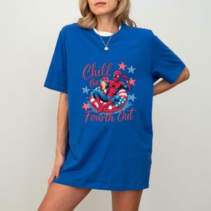 May include: A royal blue short-sleeve t-shirt featuring a graphic of Spider-Man relaxing in a red, white, and blue striped pool float while holding a drink. The text Chill the Fourth Out is printed in red, surrounded by red and white stars.