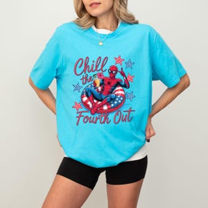 May include: A bright turquoise t-shirt featuring a graphic of Spider-Man relaxing on a red, white, and blue star-patterned pool float while holding a tropical drink. The text Chill the Fourth Out is printed in red script around the graphic.