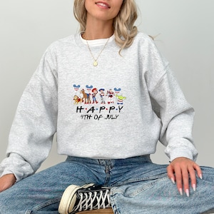 May include: A light grey crewneck sweatshirt featuring a graphic of Toy Story characters holding red, white, and blue balloons. The text below the characters reads HAPPY 4TH OF JULY in a black font. The sweatshirt has long sleeves and a ribbed collar.