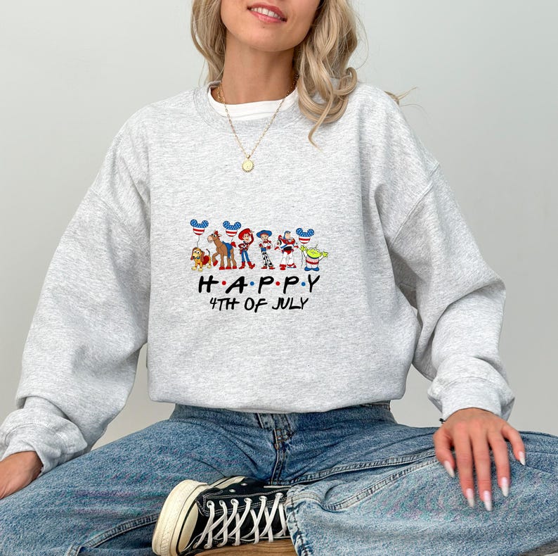 May include: A light grey crewneck sweatshirt featuring a graphic of Toy Story characters holding red, white, and blue balloons. The text below the characters reads HAPPY 4TH OF JULY in a black font. The sweatshirt has long sleeves and a ribbed collar.